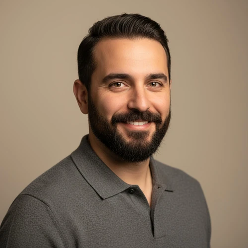 Man with dark hair and beard wearing a gray polo shirt, smiling at the camera against a plain beige background.