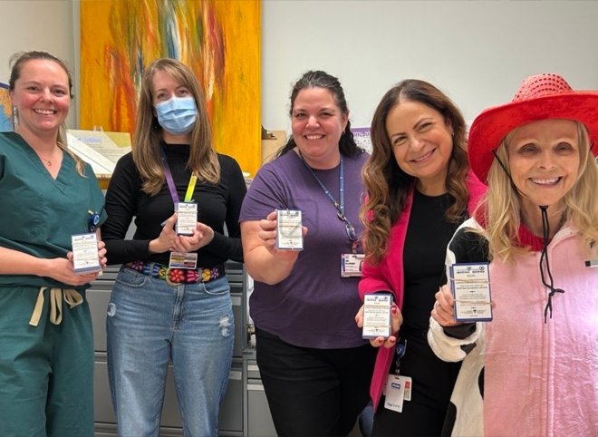 Five women stand indoors, smiling at the camera and holding up small boxed items. One woman wears medical scrubs, another wears a red hat, and one is wearing a medical mask.